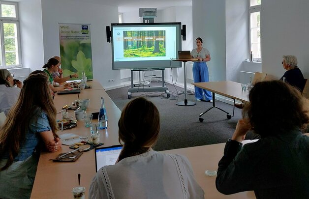 Waldwissenfrühstück 2025 Eine Gruppe von Menschen sitzt an einem Tischkreis. Eine Frau steht vor einer Leinwand und hält einen Vortrag.